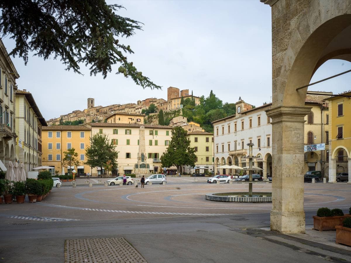 A broad Tuscan town square with café umbrellas, a fountain and ochre buildings