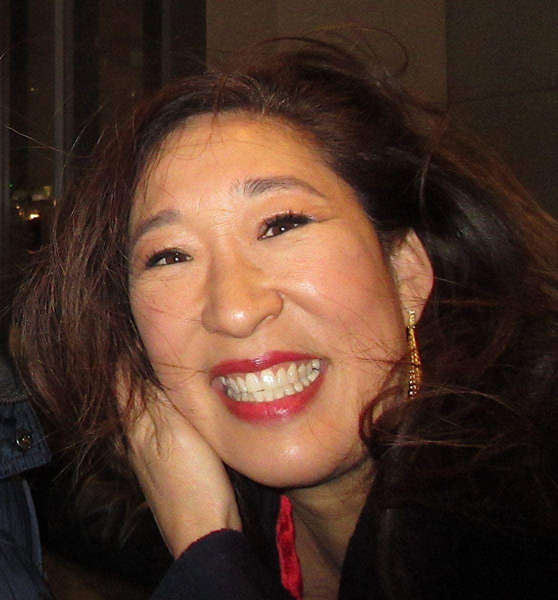 Sandra Oh smiling, dark hair loose, in a dark top against a city evening