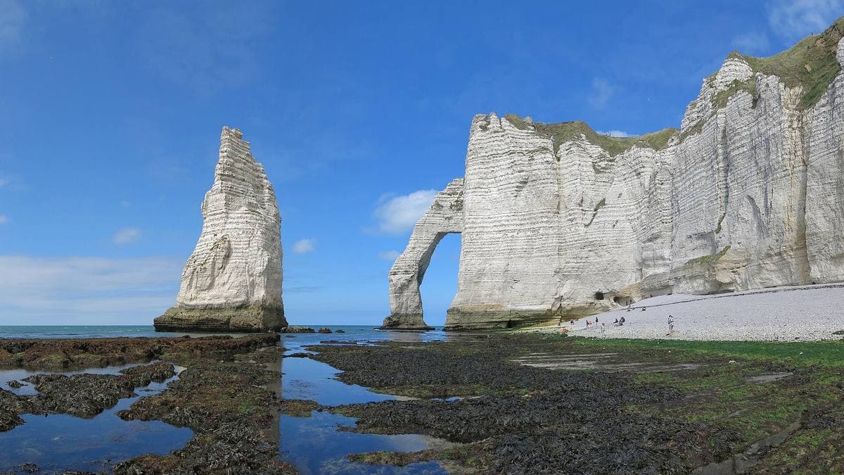 A tall chalk needle in the sea beside an arched white cliff, rocks exposed at low tide