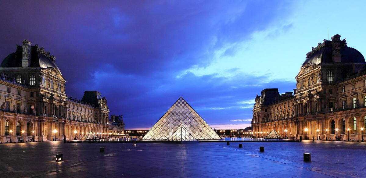 The Louvre pyramid lit up in a stone courtyard under a deep blue evening sky