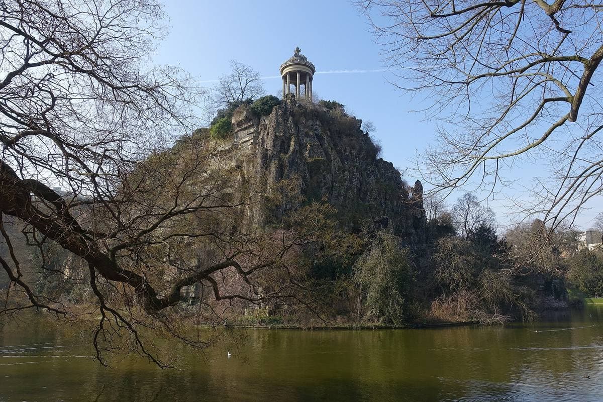 A small round temple on top of a tall rock above a lake, framed by bare branches