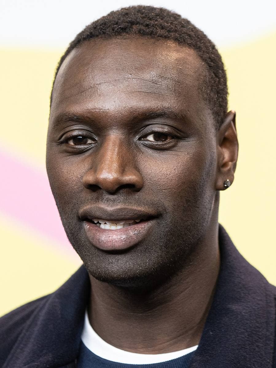 Omar Sy in a dark jacket, smiling slightly, against a pale yellow backdrop