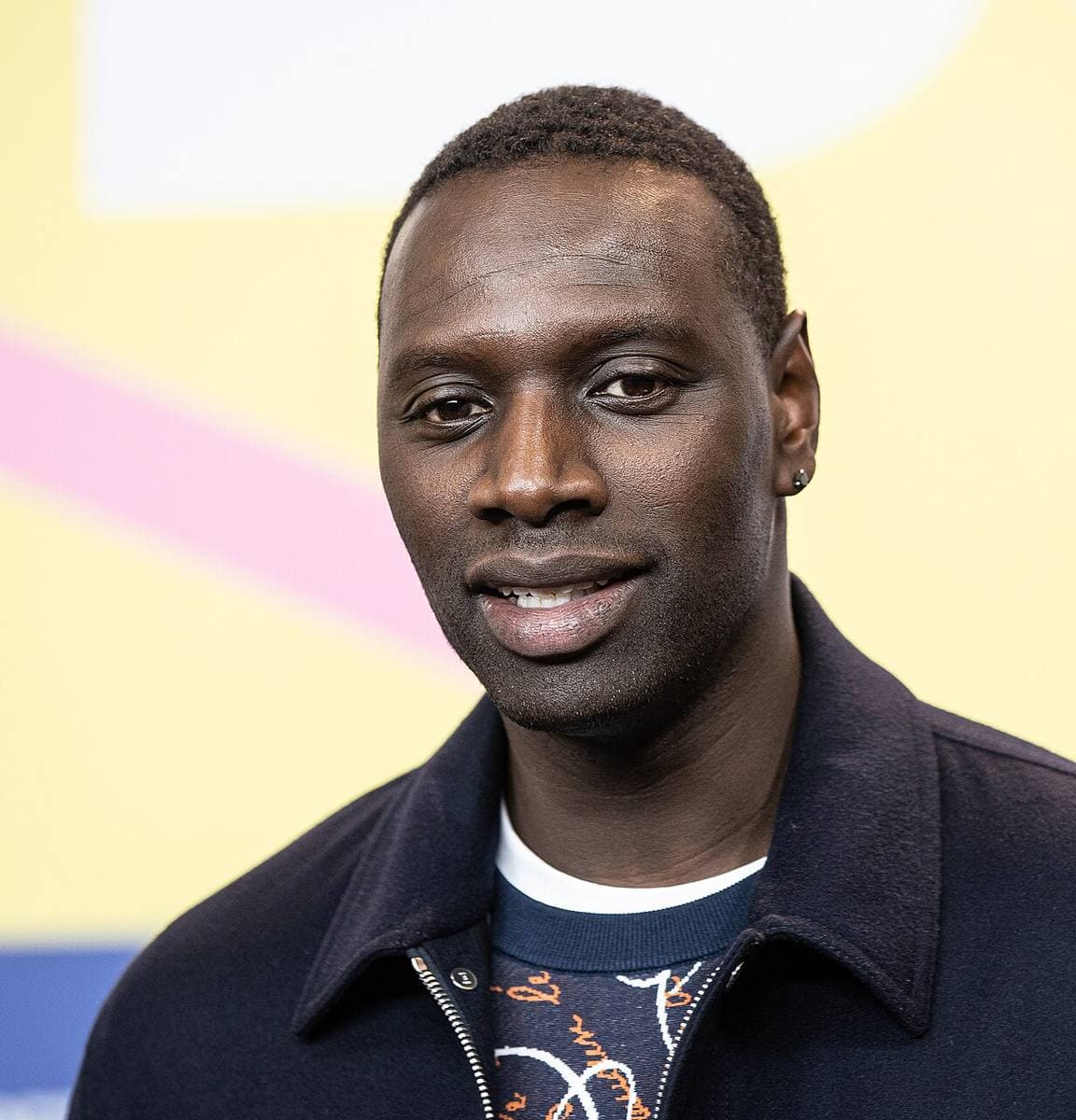 Omar Sy in a navy jacket, mid-sentence, at a press call