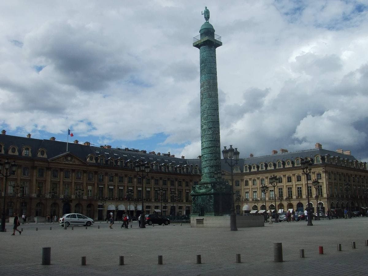 A tall bronze-green column in a square of stone façades under a cloudy sky