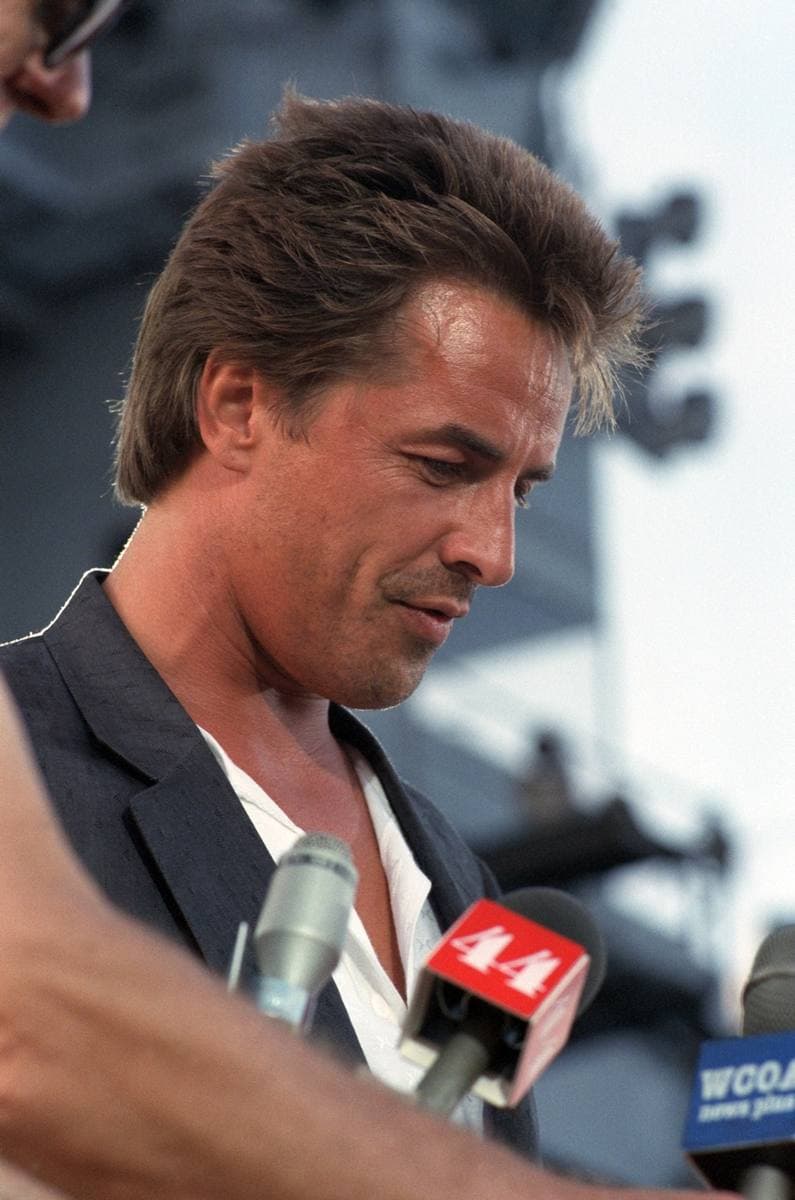 Don Johnson in a dark jacket over an open white shirt, looking down at the microphones