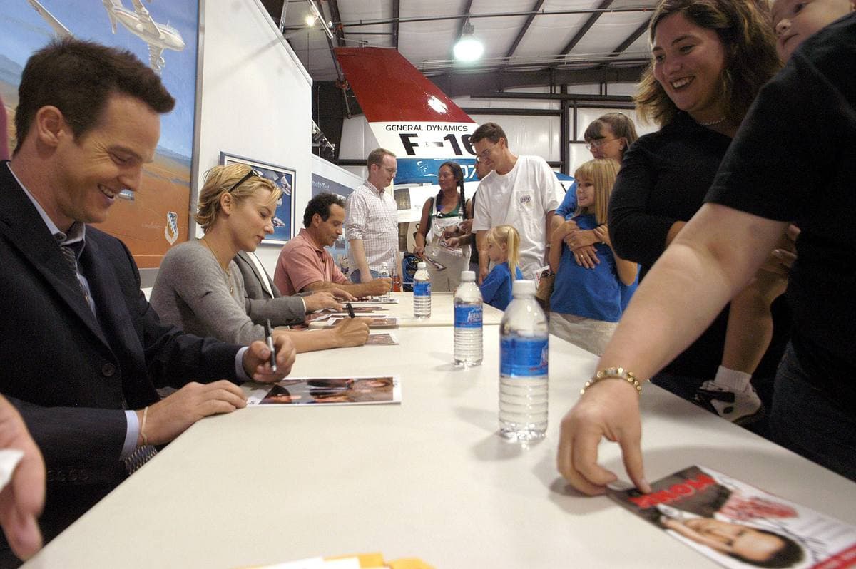 Four actors seated at a long table signing photographs for a queue of people