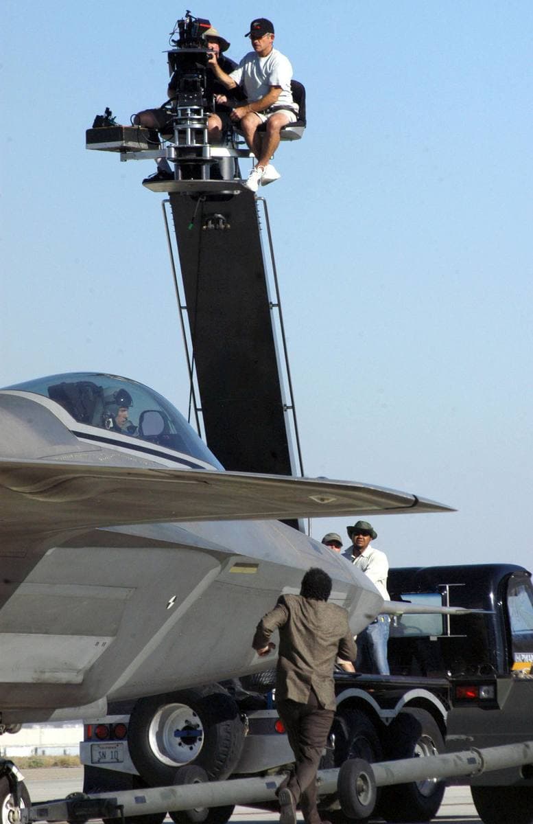 A film camera crew on a tall crane above a fighter jet, with an actor running beside it