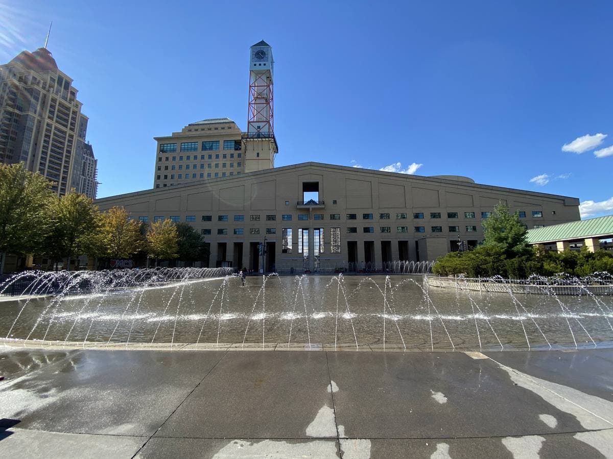 A large pale civic building with a clock tower behind a plaza of fountains