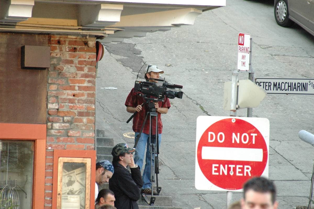 A camera operator on a tripod beside a Do Not Enter sign and a Peter Macchiarini street sign
