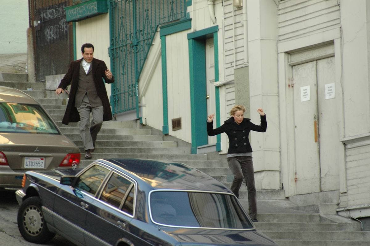 Tony Shalhoub in a long brown coat and Traylor Howard running down concrete steps between parked cars