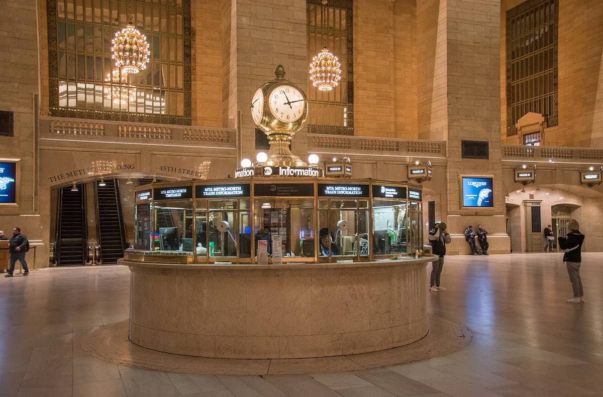 A round booth with a brass four-faced clock in a vast marble concourse