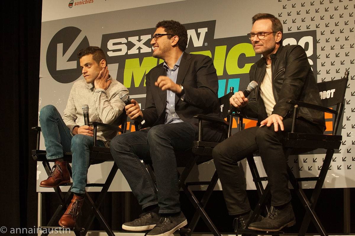 Rami Malek, Sam Esmail and Christian Slater seated on a festival stage with microphones