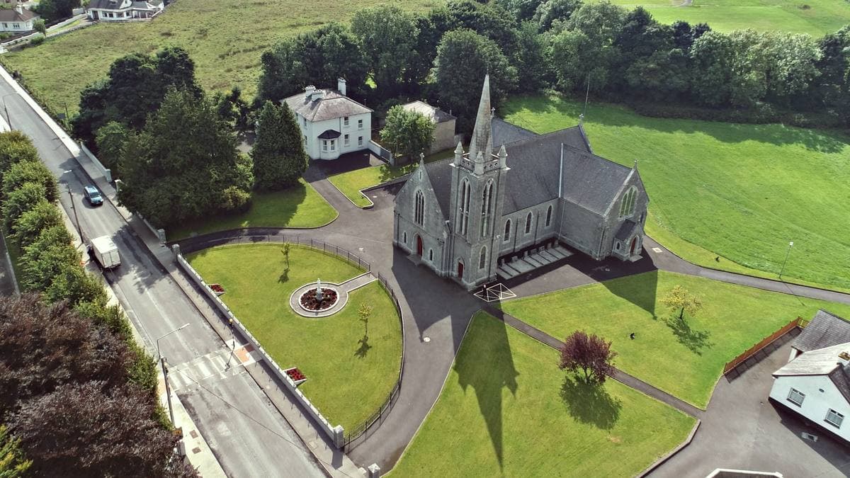 An aerial view of a grey stone church with a spire, lawns around it and a road alongside