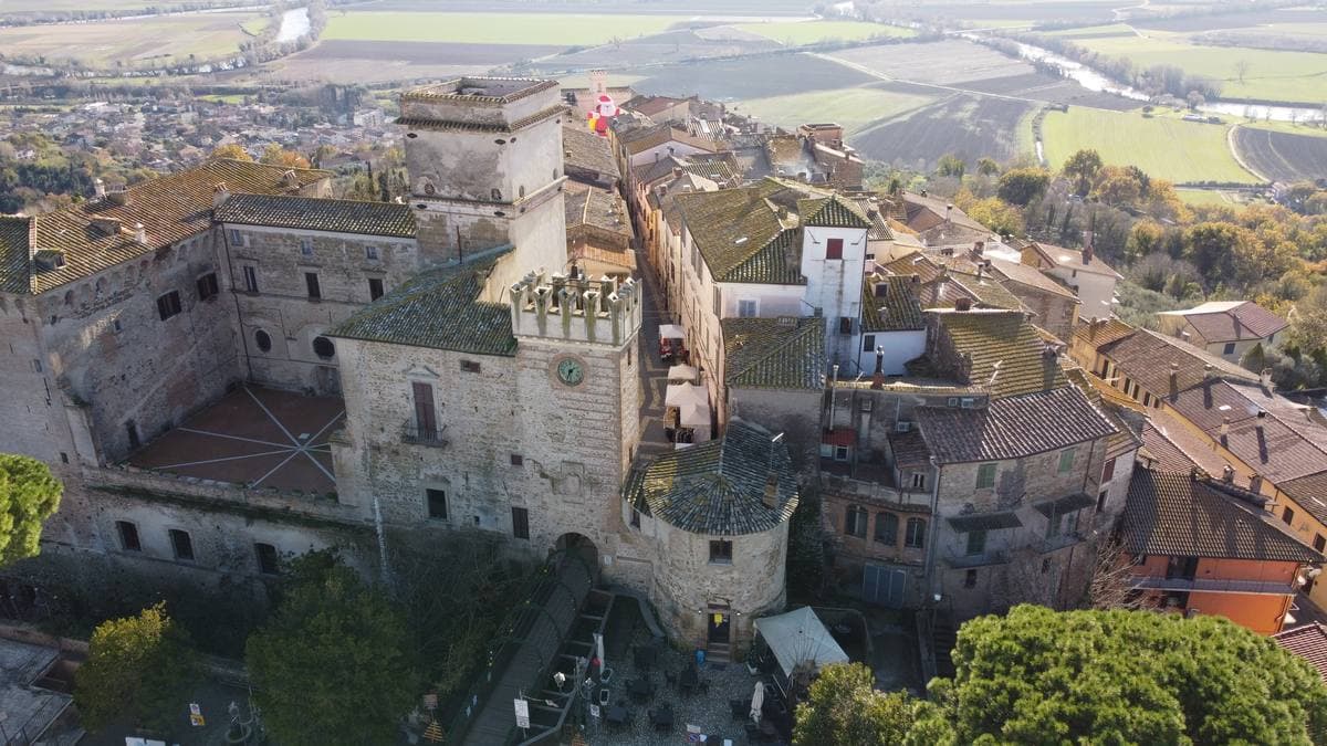 An aerial view of a hilltop Italian town of tiled roofs and a castle tower, with fields beyond