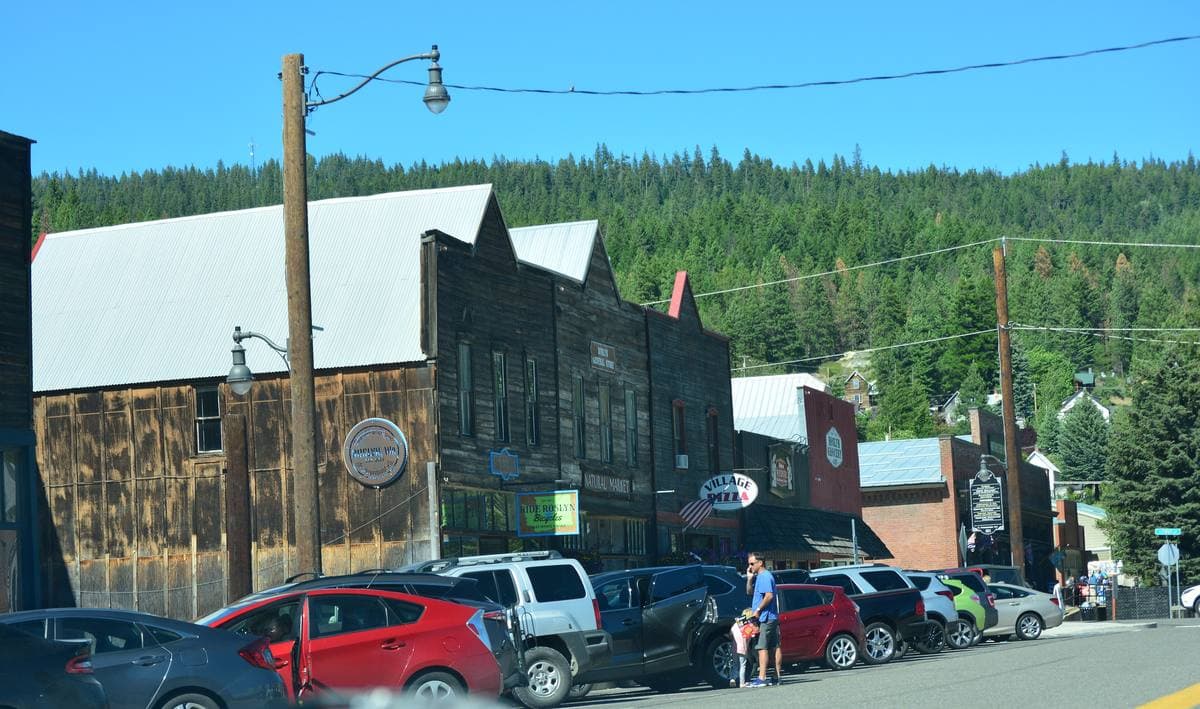 A row of old wooden false-front shops on a small-town street with cars parked and forest behind