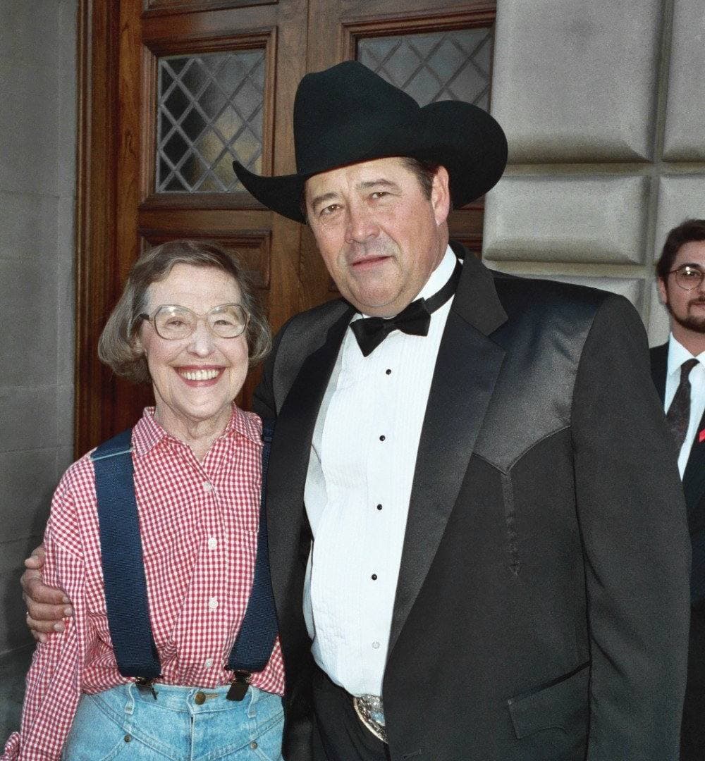An older woman in a gingham shirt, braces and jeans beside a tall man in a black cowboy hat and dinner jacket