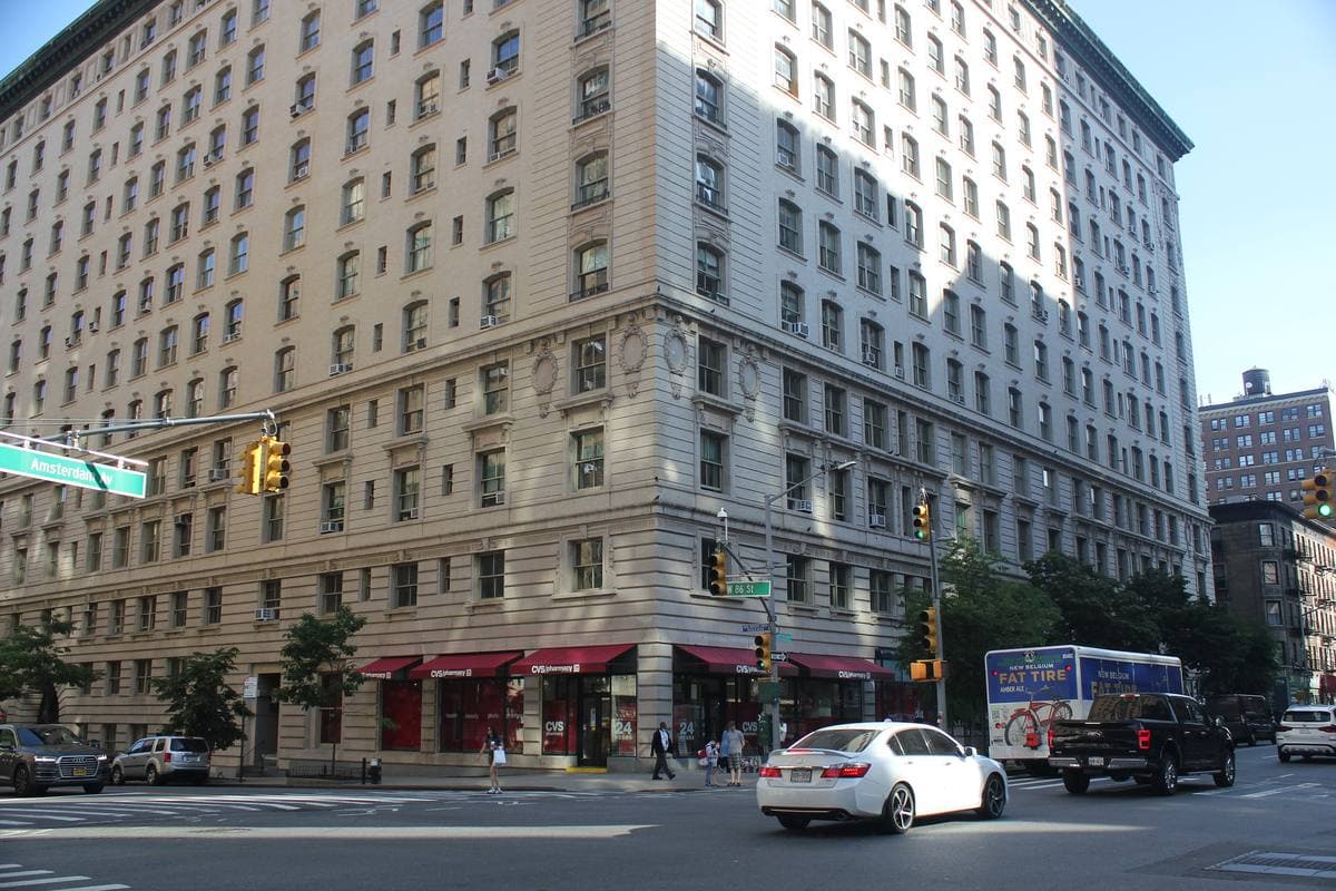 A thirteen-storey limestone apartment block filling a city corner, with shops at street level and traffic on Broadway
