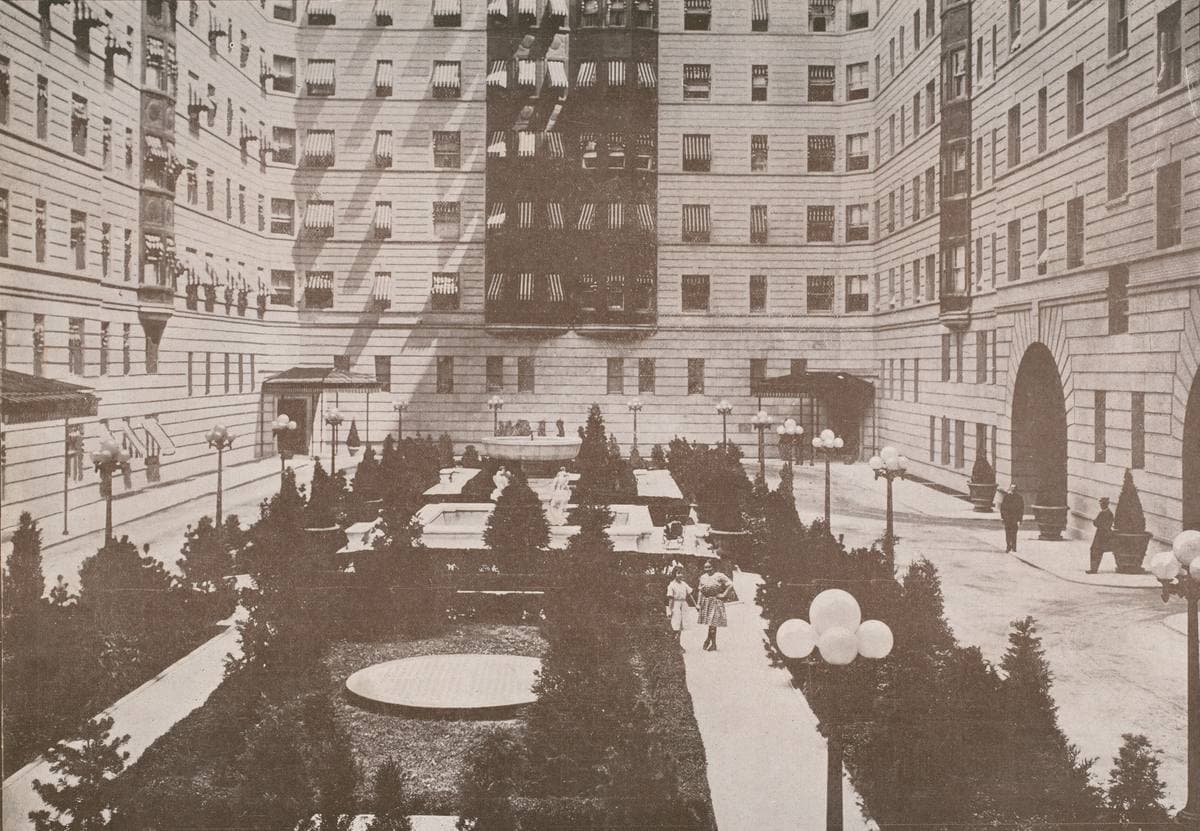 A sepia photograph of a vast courtyard ringed by tall apartment walls, with formal lawns, clipped trees, round lamps and a carriage drive