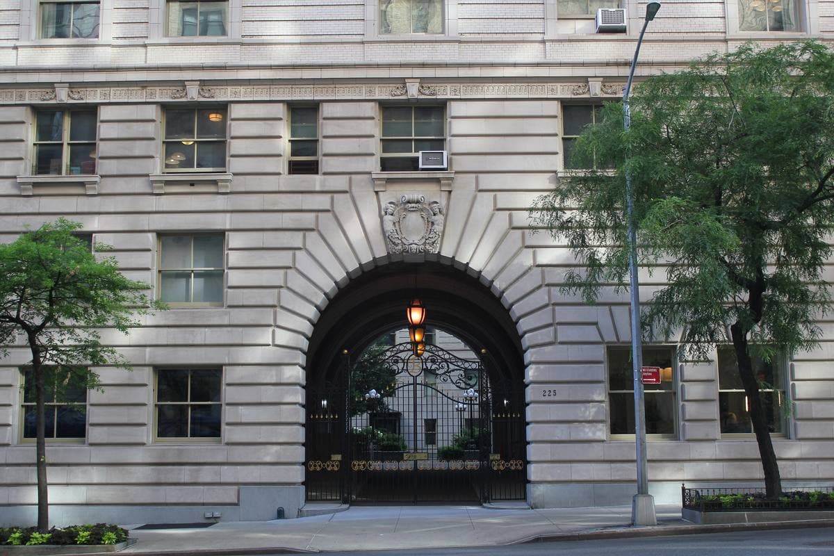 A tall rusticated stone arch with wrought-iron gates and a lantern, set into a pale limestone facade between two street trees