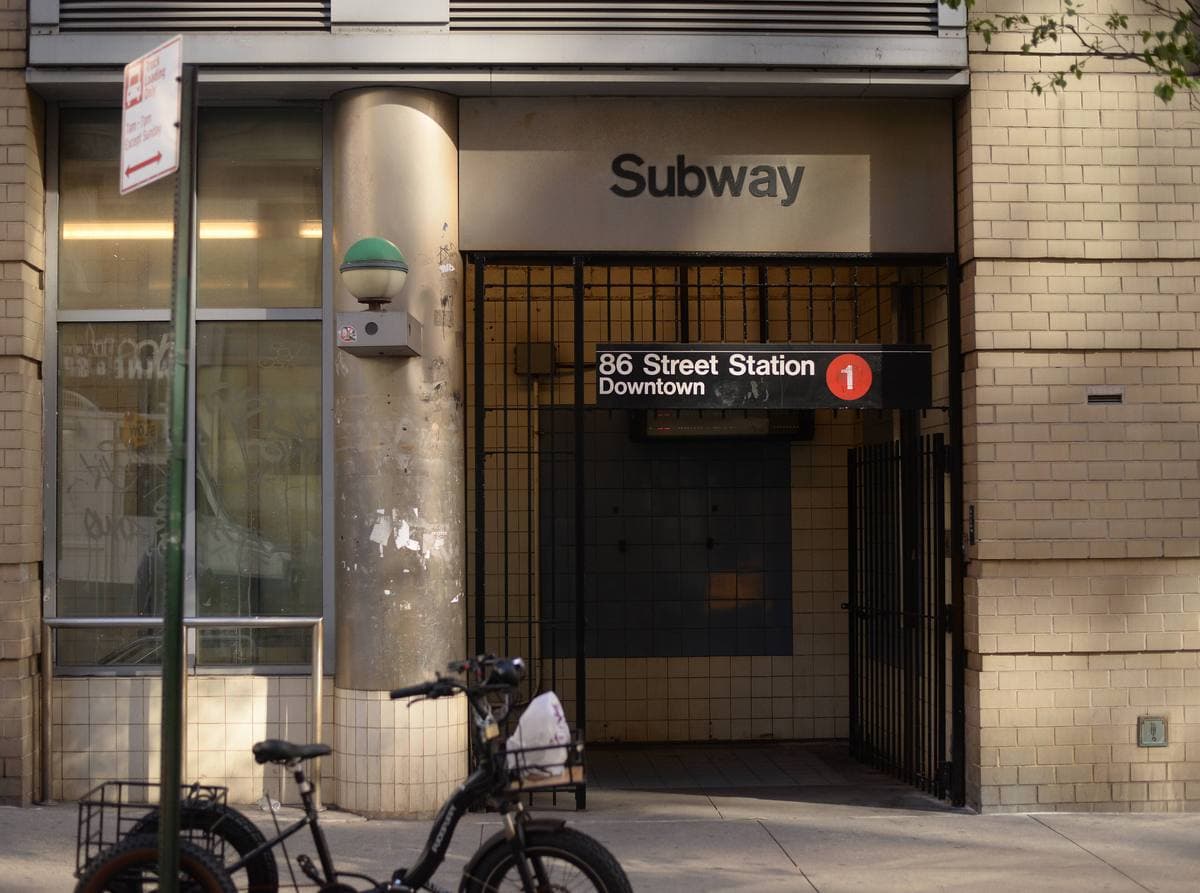 A subway entrance set into a building, with a sign for the 86 Street station and the red 1 train roundel, and a bicycle outside
