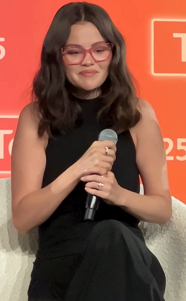 A young woman with long dark hair and red glasses, smiling and holding a microphone