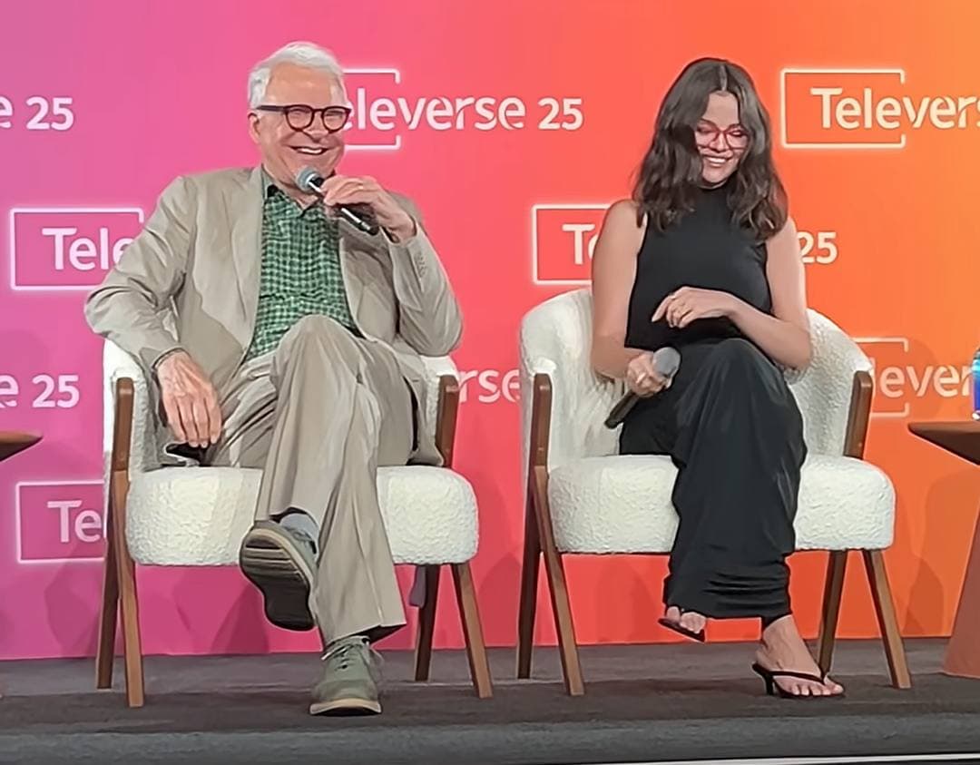 An older man in glasses and a beige suit laughing into a microphone beside a young woman in black and red glasses, both seated in white armchairs