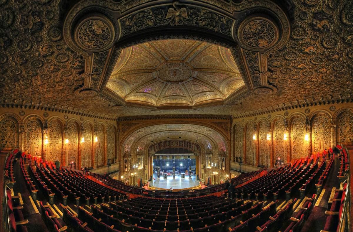 A wide view of an ornate gilded auditorium with a carved ceiling dome, red seats and a lit stage