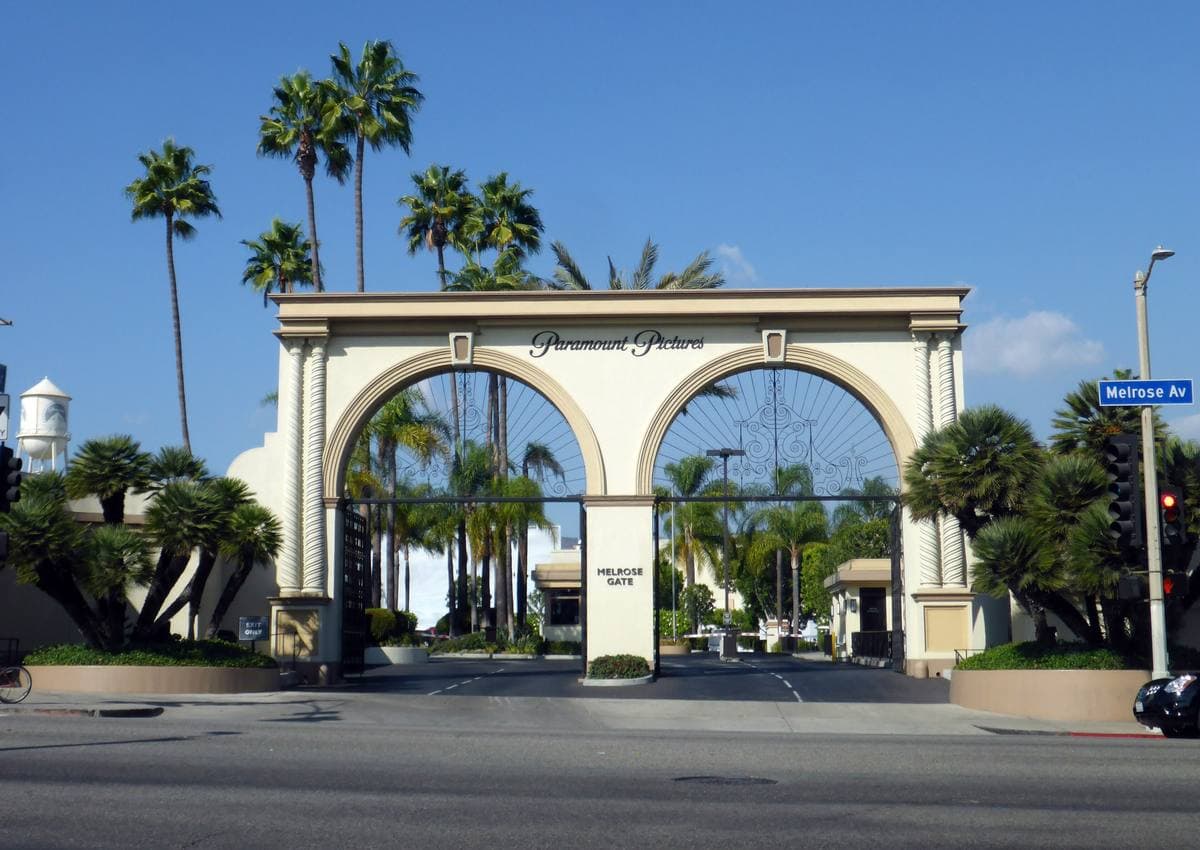 A cream double-arched studio gateway with iron gates, palm trees behind it and a water tower in the distance