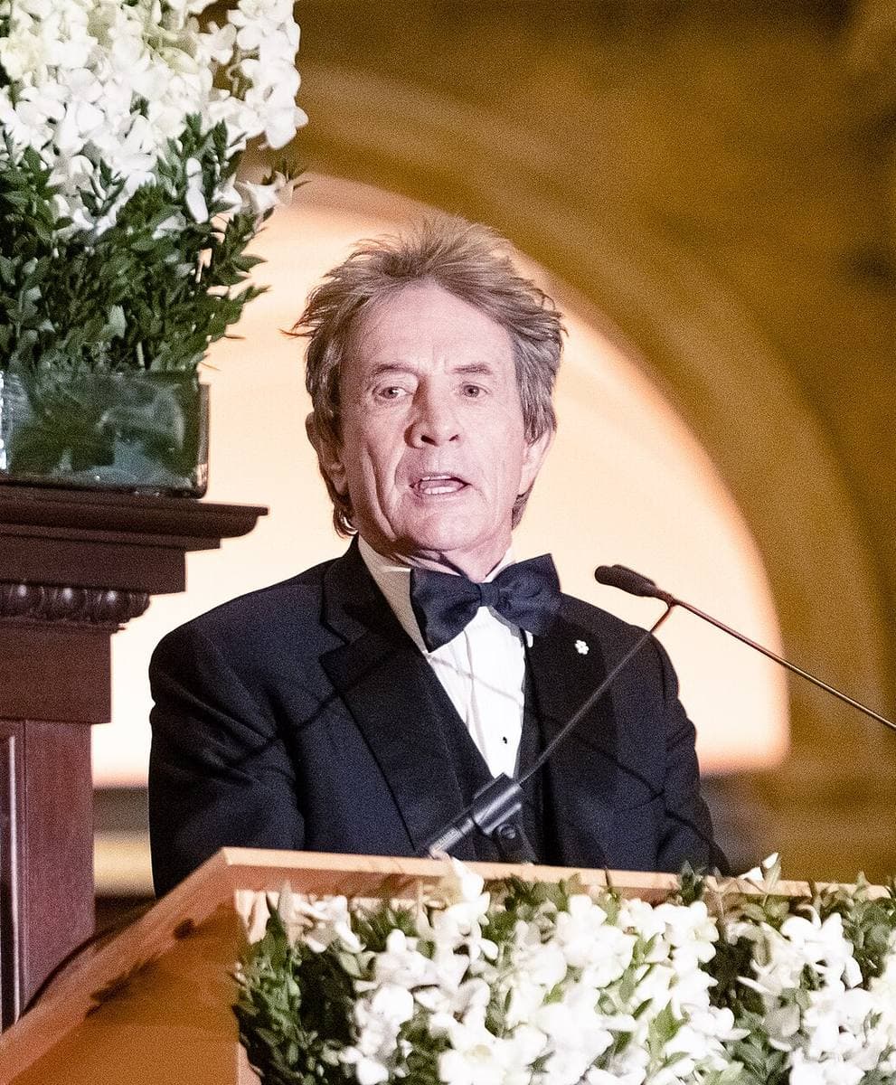 A grey-haired man in a dinner jacket and bow tie speaking at a lectern decked with white flowers