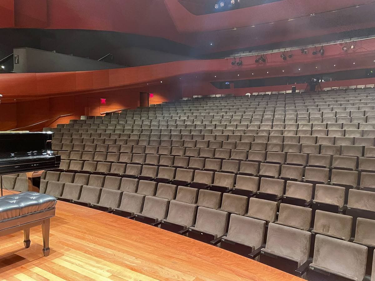 Rows of grey concert seats rising from a wooden stage with a piano, under warm wood-panelled walls