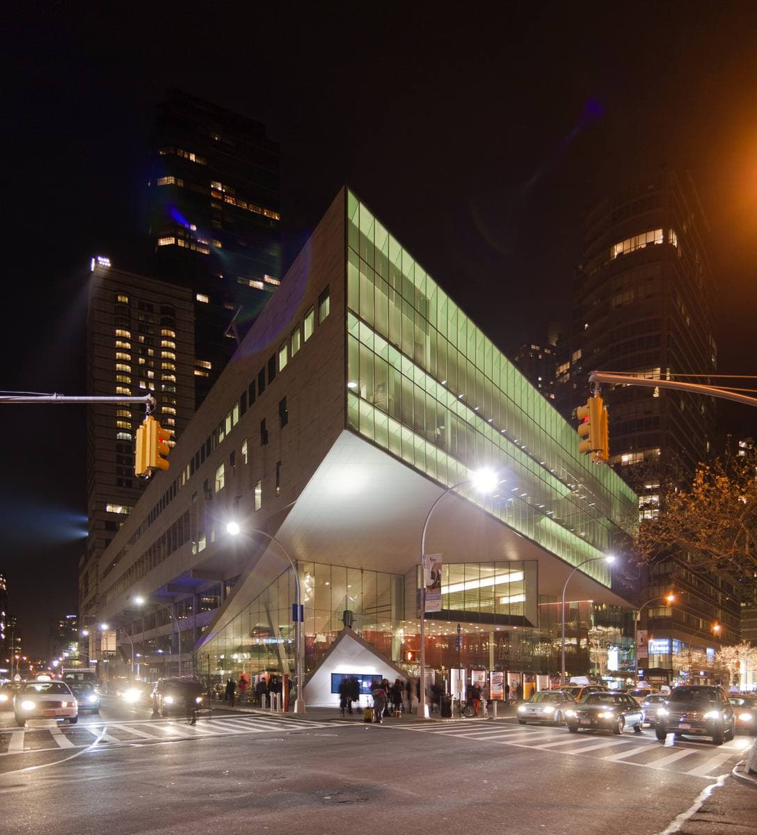 A modern concert hall with a sharply cantilevered glass corner glowing at night over Broadway