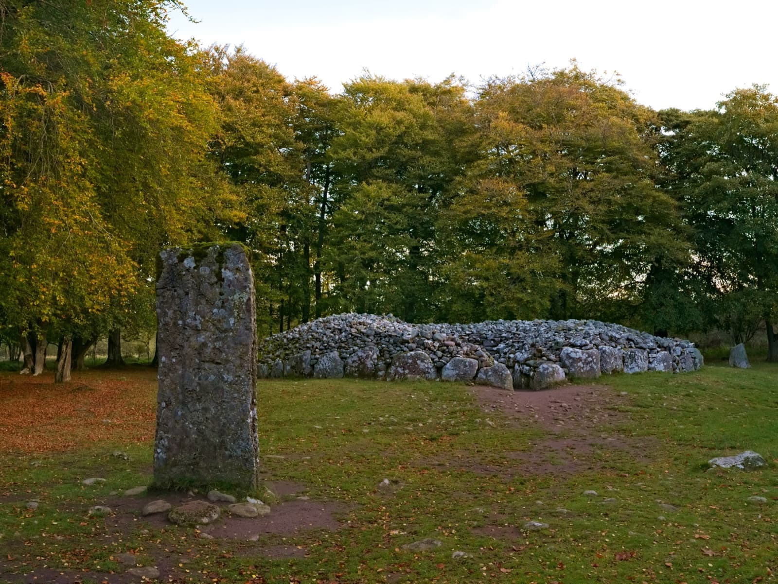 A tall standing stone in grass before a low round cairn under autumn trees