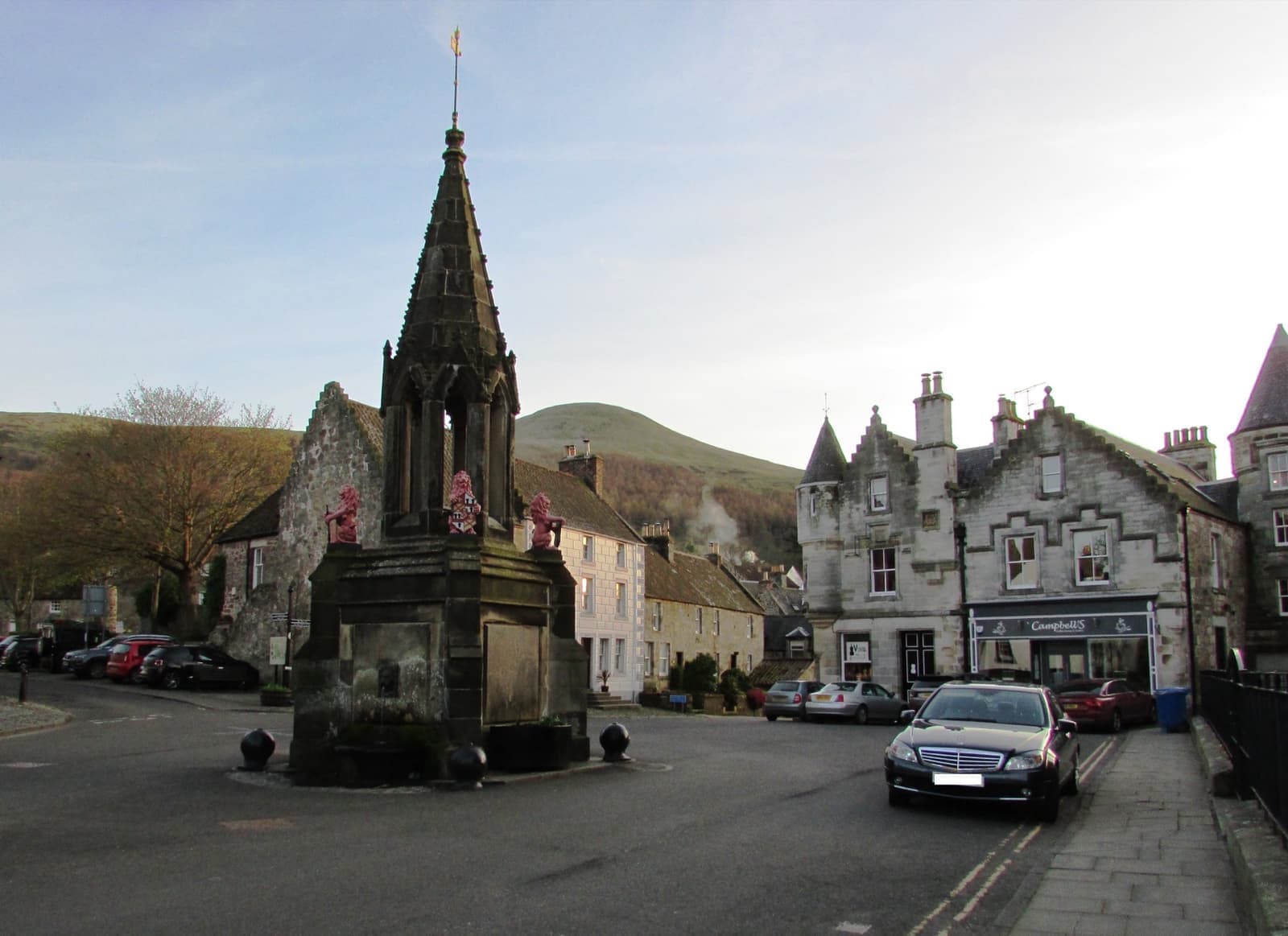A tall Gothic stone fountain in a village square with the Lomond hills behind