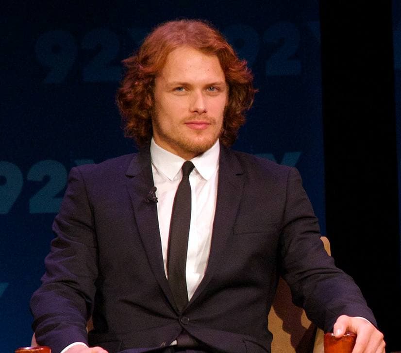 Sam Heughan with reddish curly hair and a short beard, in a dark suit and tie, seated on stage