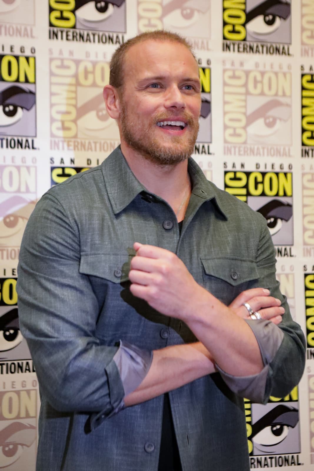 Sam Heughan in a grey-green shirt, arms folded, smiling