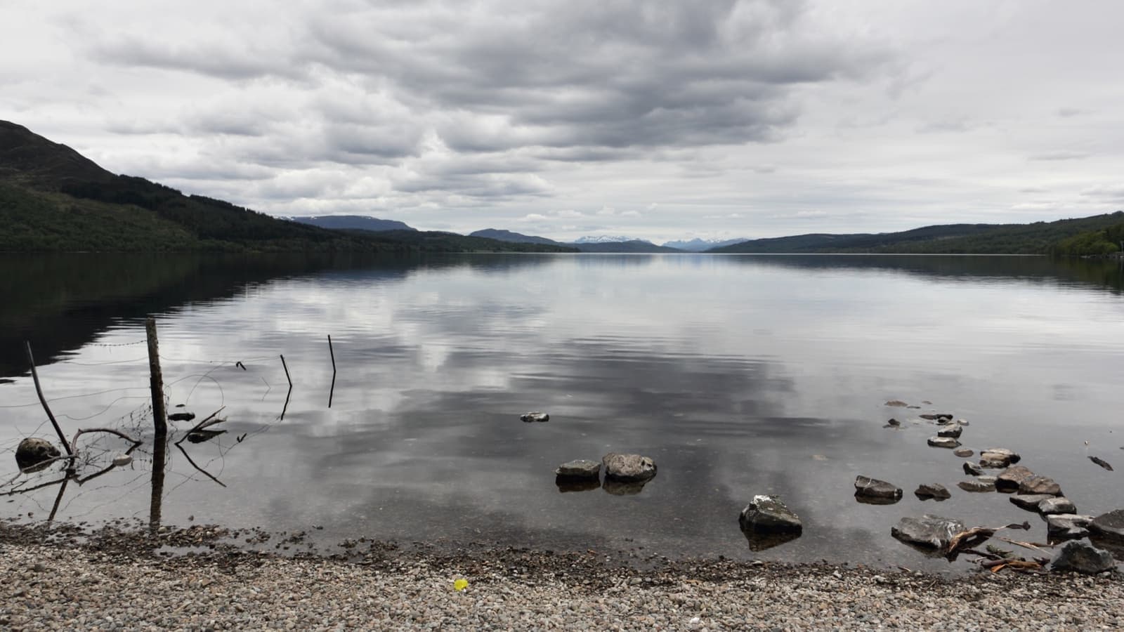 A long grey loch between low wooded hills under cloud