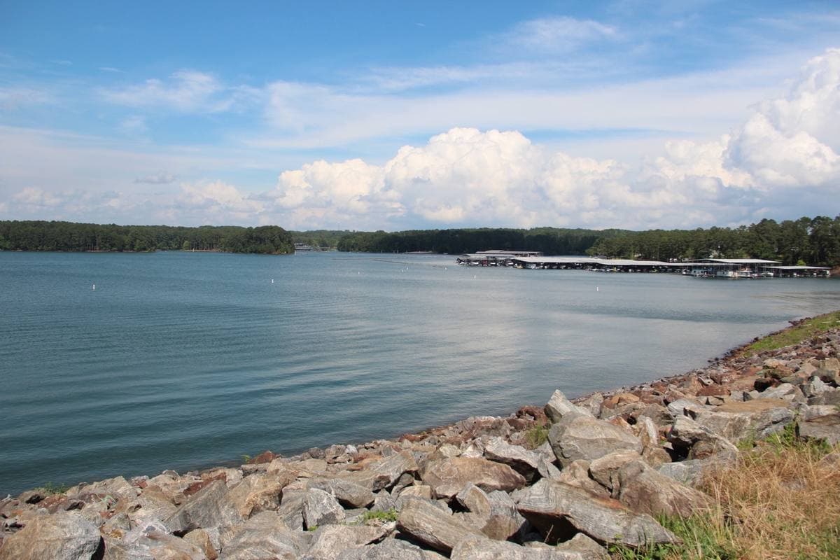 A wide lake under summer clouds, rocks in the foreground and long covered boat docks across the water