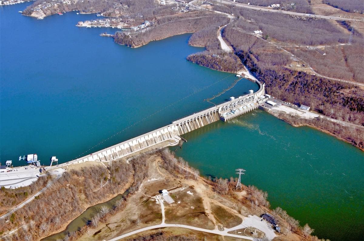 Aerial view of a long concrete dam across a blue lake between wooded hills