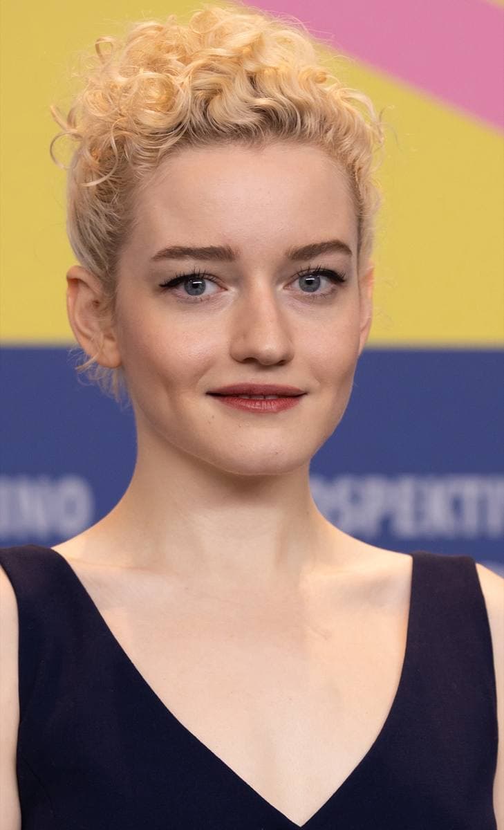 Julia Garner with curled blonde hair pinned up, in a dark top, against a yellow and blue backdrop