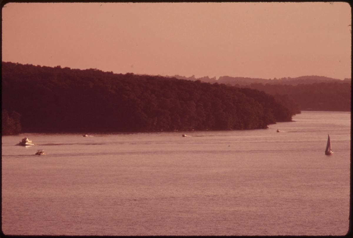 Small boats and a sailboat on a wide lake under a warm evening haze, wooded ridge behind