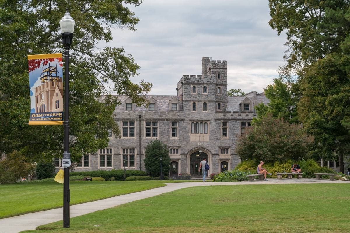 A grey stone collegiate building with battlements behind a lawn and a lamp post