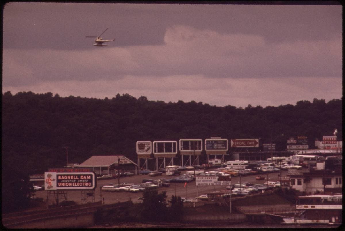 Billboards, cars and a Bagnell Dam Union Electric sign at the end of a commercial strip, wooded hills behind