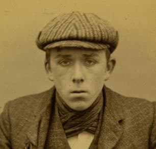 A young man in a checked flat cap and neckerchief in a sepia police photograph