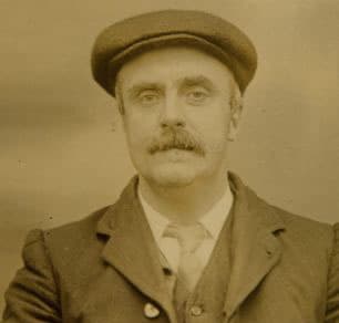A moustached man in a flat cap in a sepia police photograph