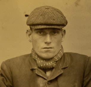 A young man in a flat cap and spotted neckerchief in a sepia police photograph