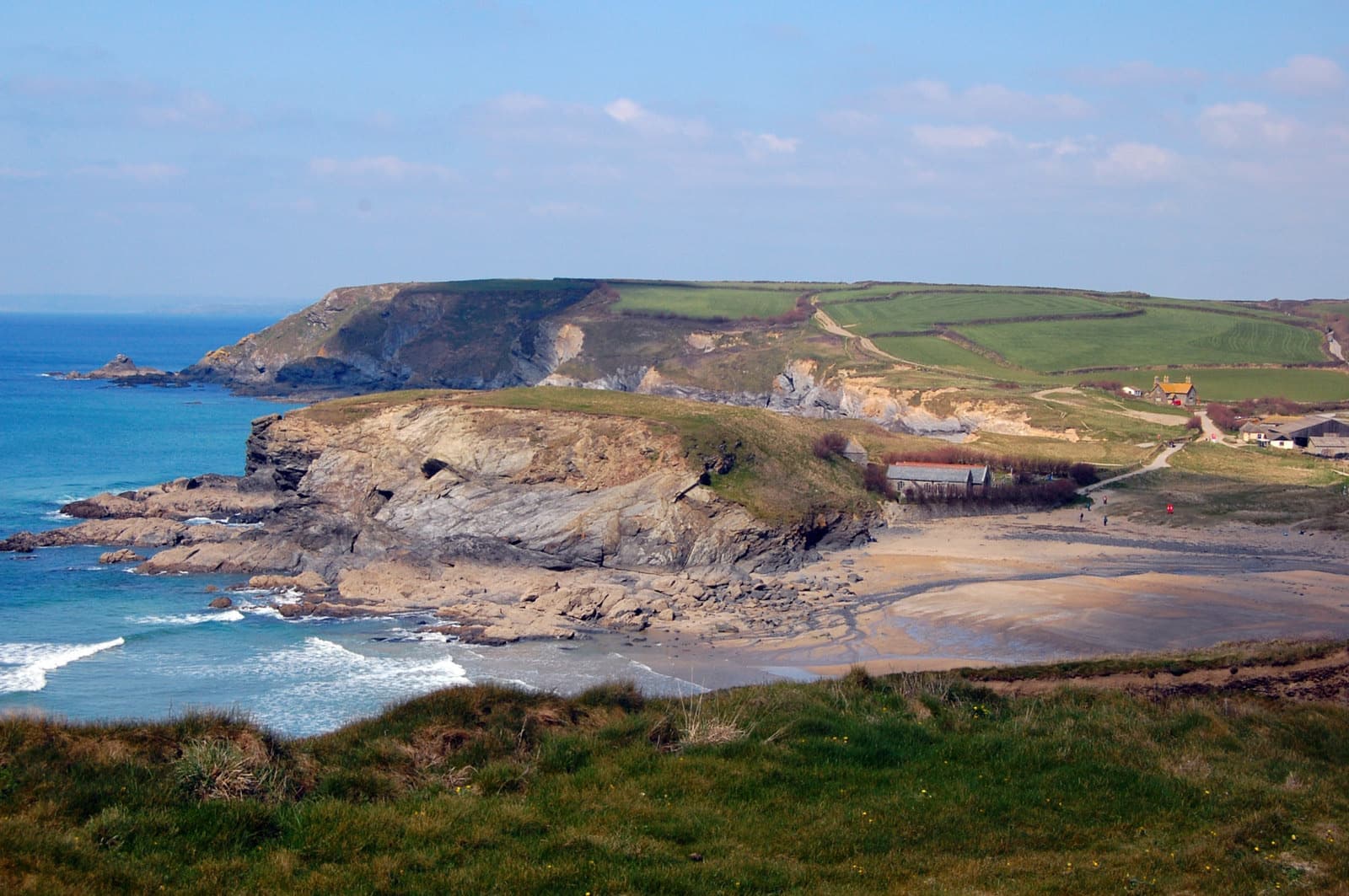 A sandy cove below a low green headland with fields beyond and blue sea in front