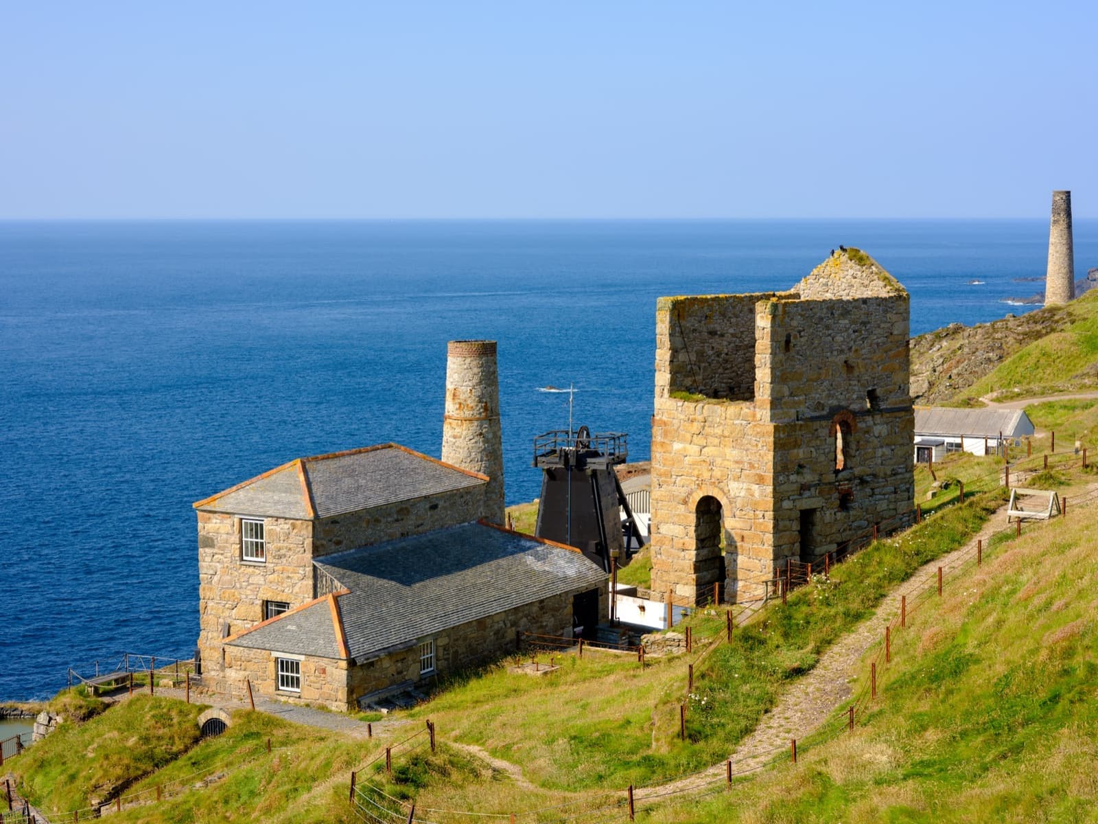 Granite engine houses with a black headframe and a tall chimney on a green clifftop over the sea