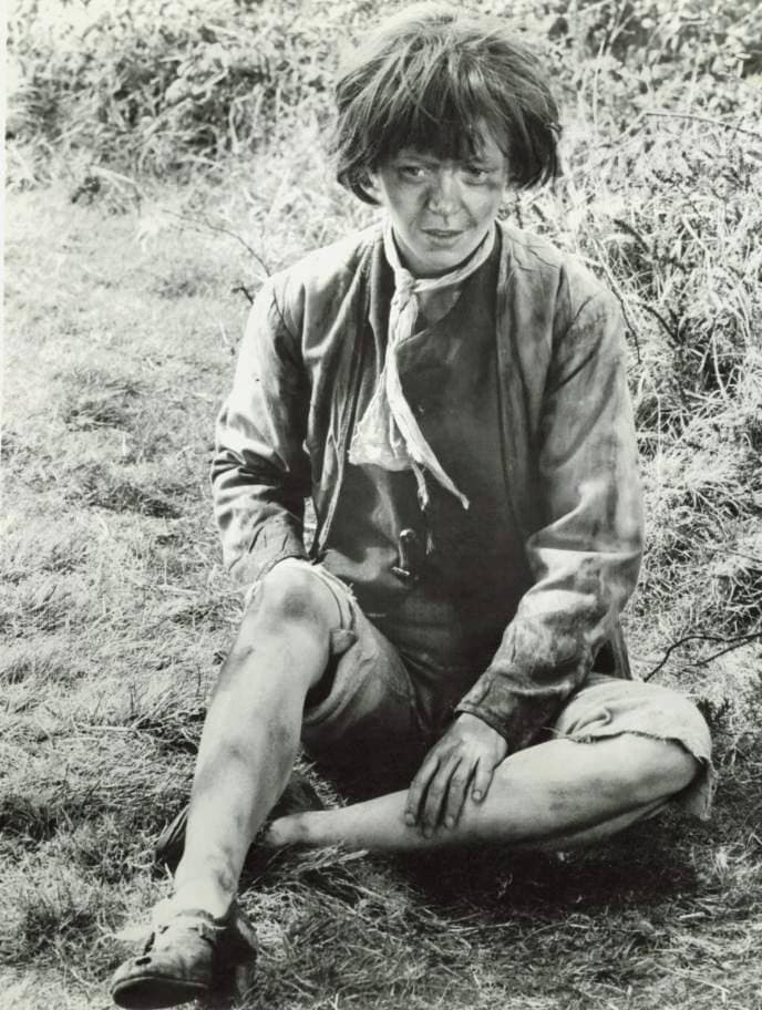 Angharad Rees in costume as a ragged girl, sitting barefoot in long grass