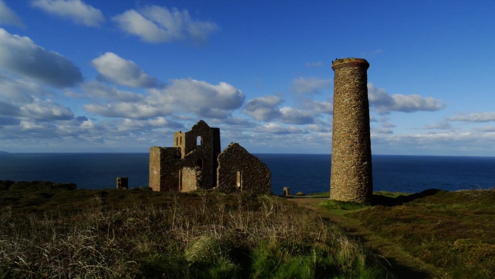 A roofless engine house and a round chimney on heathland against a blue sky and the sea