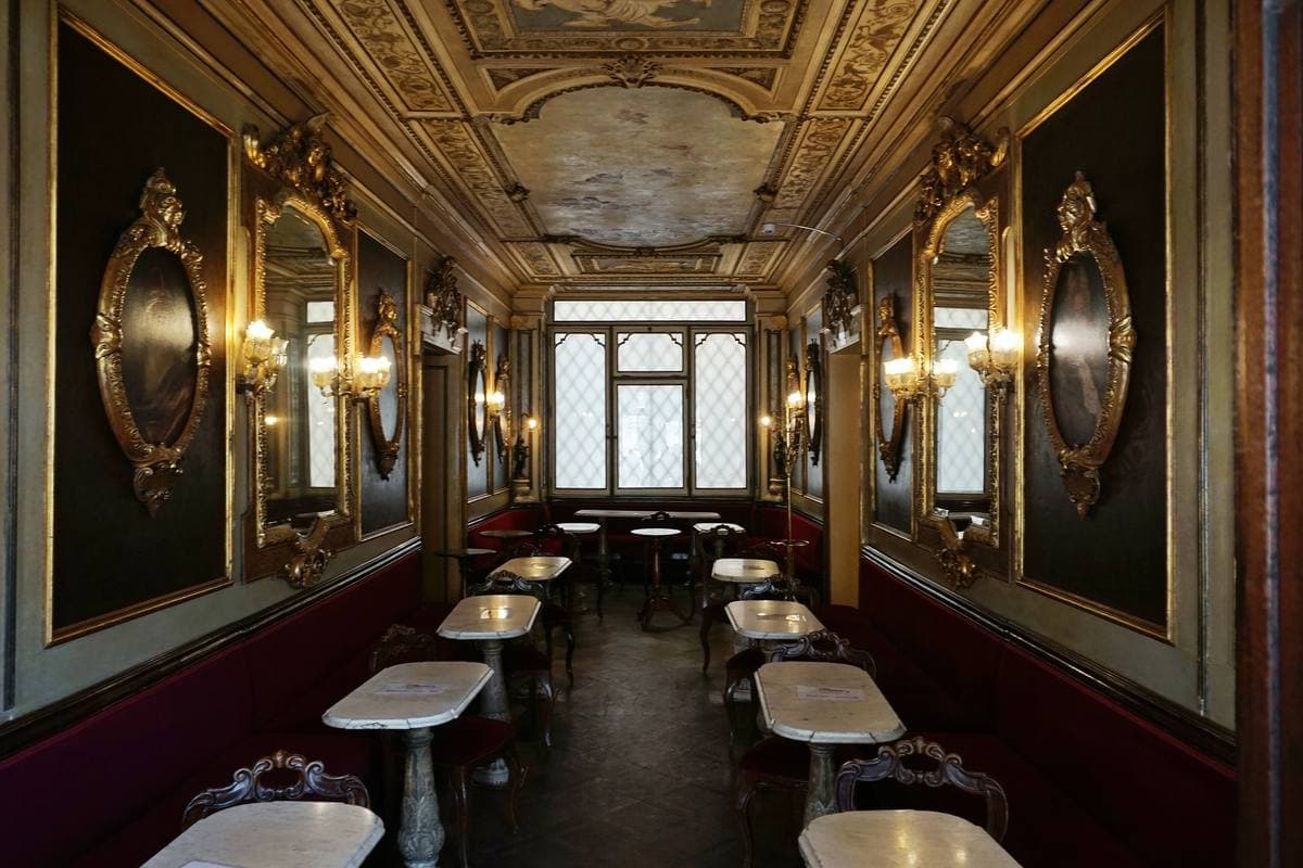 A narrow café room of mirrors, painted panels and small marble tables